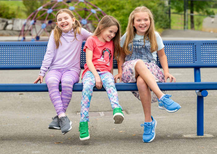 Three young girls sitting and laughing on a blue bench at a playground, wearing colorful outfits and adaptive sneakers, enjoying a playful outdoor moment.