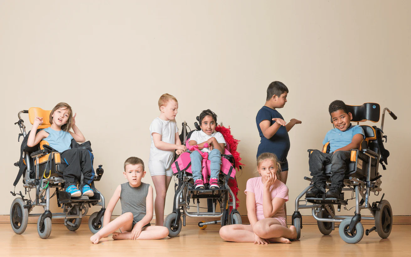 A diverse group of children, including those in wheelchairs and seated on the floor, pose together against a plain wall, wearing adaptive clothing designed for comfort and accessibility.