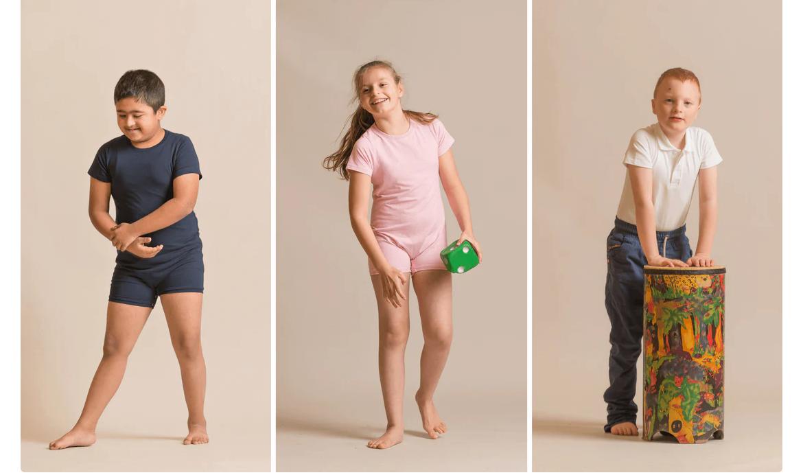 Three children wearing KayCey adaptive clothing, including bodysuits and polo shirt, standing and smiling in a studio setting.
